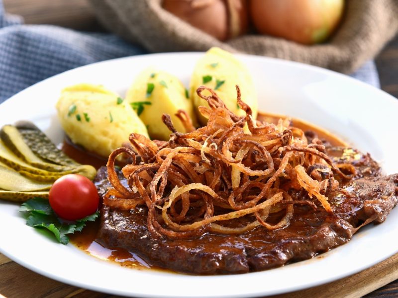 Ein Zwiebelrostbraten mit Sauce und Beilagen auf einem hellem Teller aus Porzellan. / Draufsicht, schräg.