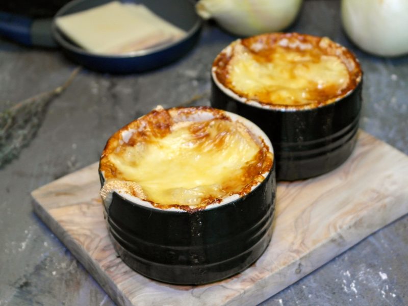Zwei schwarze Schälchen mit der überbackenen Zwiebelsuppe auf einem kleinen Holzbrett und Steinuntergrund. Alles von schräg oben fotografiert.