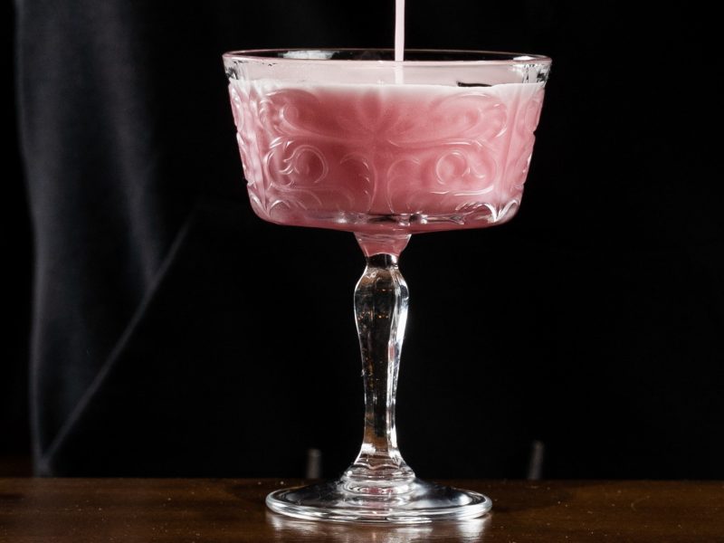 Pink Gin Sour wird in ein elegantes, verziertes Cocktailglas aus Glas mit filigranem Stiel auf dunklem Hintergrund gegossen.