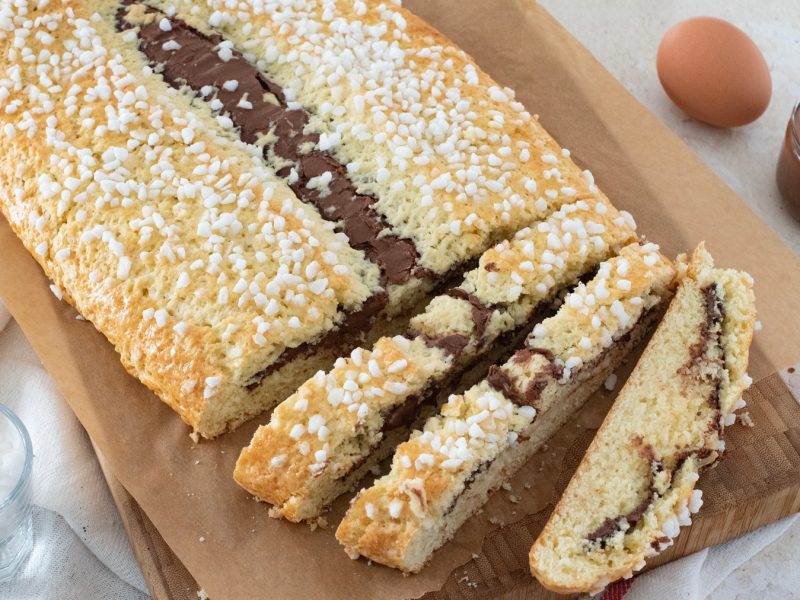 Frisch gebackener Bensone mit glänzender Schokofüllung und knusprigem Hagelzucker auf einem Holzbrett, umgeben von Zutaten.