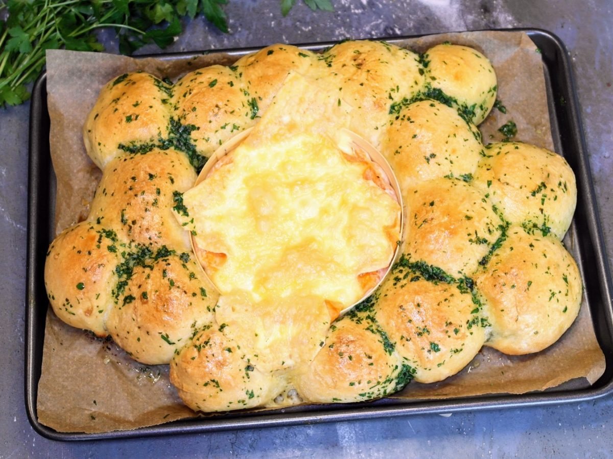 Ein Blech mit dem Brötchenkranz und Ofenkäse auf einem steinernen Tisch von schräg oben fotografiert. Im Hintergrund unscharf ein Bund Petersilie erkennbar.