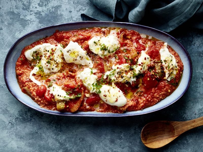 Ovaler Teller mit Büffelmozzarella auf Röstgemüse à la Romesco, daneben ein Holzlöffel auf blaugrauem Untergrund.