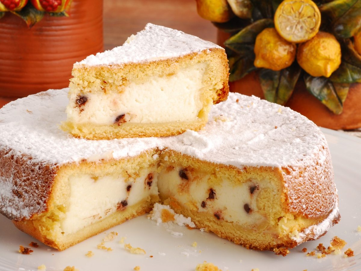 Ein angeschnittene Cassata al forno mit Schokoladenstückchen und Puderzucker steht auf einem weißen Teller, im Hintergrund dekorative Keramiktöpfe.