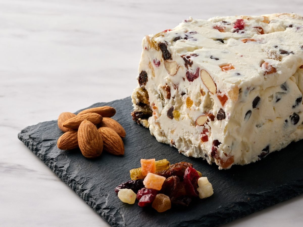 Ein Stück cremiges Cassata-Semifreddo mit Mandeln und Trockenfrüchten liegt auf einer schwarzen Schieferplatte, daneben Mandeln und bunte kandierte Früchte.
