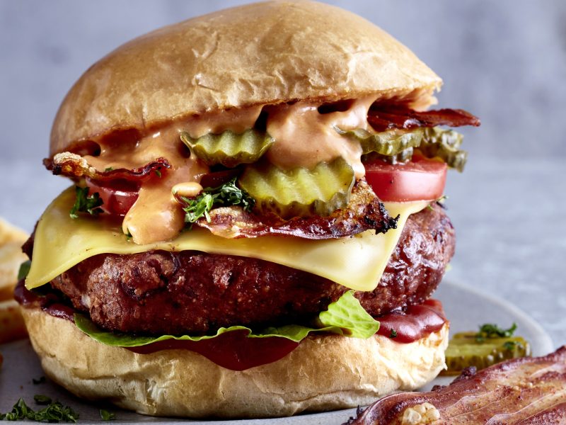 Cheesy Böhnchenburger mit Pommes vor grauem Hintergrund, dazu Pommes und veganer Bacon.