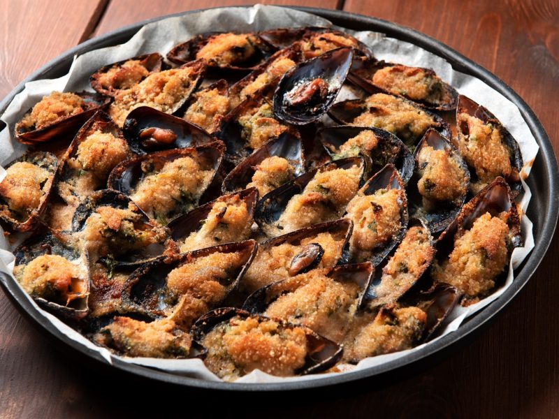 Nahaufnahme von knusprigen Cozze gratinate mit goldener Panade und Kräutern, angerichtet auf Backpapier in schwarzer Form.
