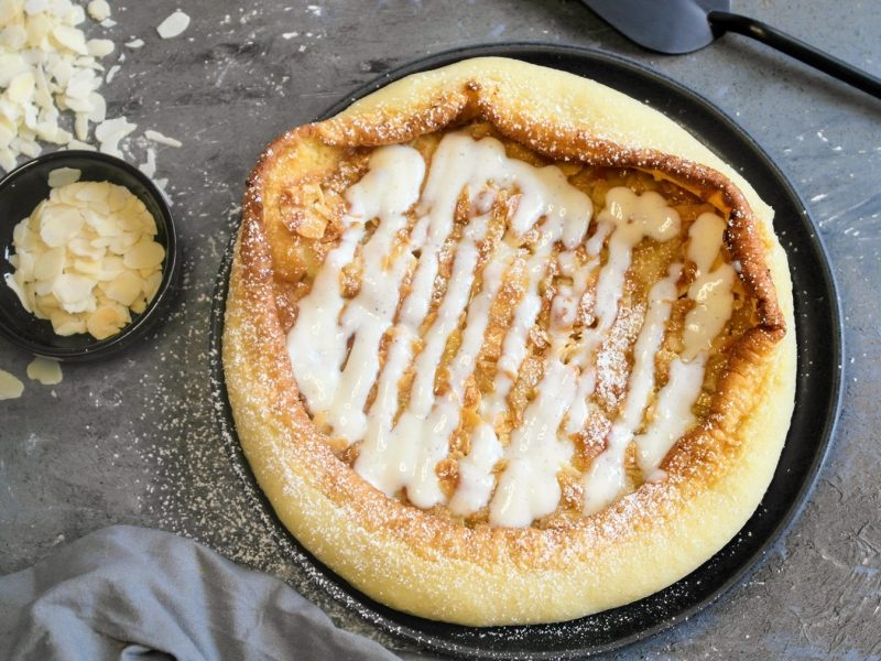 Ein schwarzer Teller mit dem Bienenstich-Pfannkuchen auf steinernem Untergrund, mit einem grauen Tuch und einem Schälchen mit Mandelblättchen.