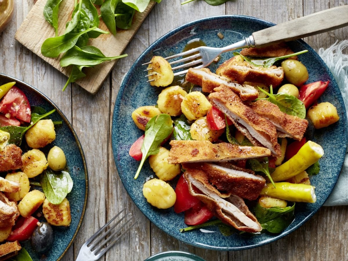 Antipasti-Schnitzel-Salat auf zwei dunkelblauen Tellern verteilt auf hölzernem Hintergrund.