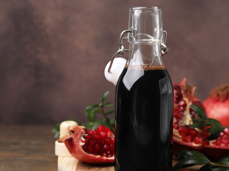Frontal: Eine Flasche mit einem selbst gemachten Balsamico-Dressing mit Granatapfel vor einem dunklen Hintergrund. Daneben liegen Teile eines Granatapfels.