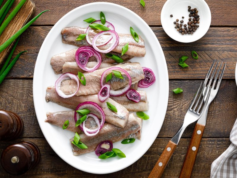 Draufsicht: Mehrere Fischfilets, beziehungsweise Bismarckheringe, auf einem hellen Teller. Der steht auf einem rustikalen Holztisch. Drum herum liegen Bestecke, Frühlingszwiebeln, Gewürzmühlen und ein Tuch.