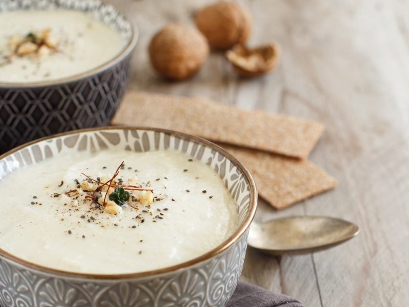 Draufsicht, schräg: Zwei Schüsseln mit einer Blumenkohlsuppe mit Walnuss auf einem rustikalen Untergrund aus Holz. Drum herum liegen ein silberner Löffel und Walnüsse.