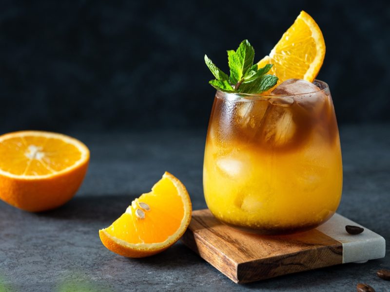Frontal: Ein Glas mit einem Getränk aus Kaffee und Orangensaft in einem Glas. Es ist mit Minzblättern und einer halben Scheibe Orangensaft verzieret. Daneben liegen eine halbe und eine viertel Orange.