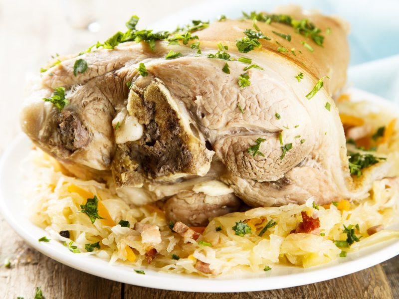 Frontal: Ein heller Teller aus Porzellan mit einem Eisbein mit Sauerkraut, dass mit frischer Petersilie bestreut wurde.