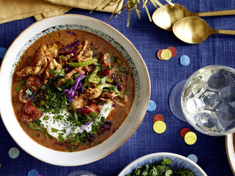 Hähnchensuppe Döner-Art in einer Schüssel auf dunkelblauem Hintergrund. Rechts oben goldene Löffel und Konfetti auf dem Hintergrund verteilt.