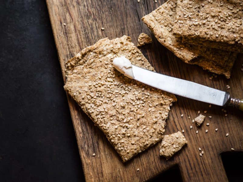 Draufsicht: Dunkler Untergrund, Holzbrettchen, zwei Scheiben Knäckebrot, ein Küchenmesser mit Butter,