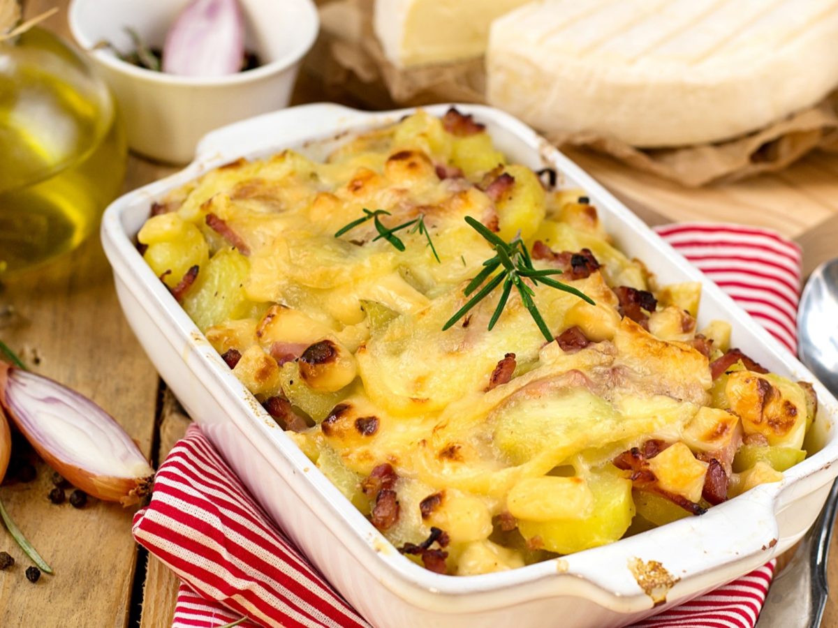 Draufsicht, schräg: Eine helle Form mit einem Kartoffelauflauf mit Schinken steht auf einem Untergrund aus Holz. Drum herum liegen Zwiebeln, ein Löffel und ein Tuch.