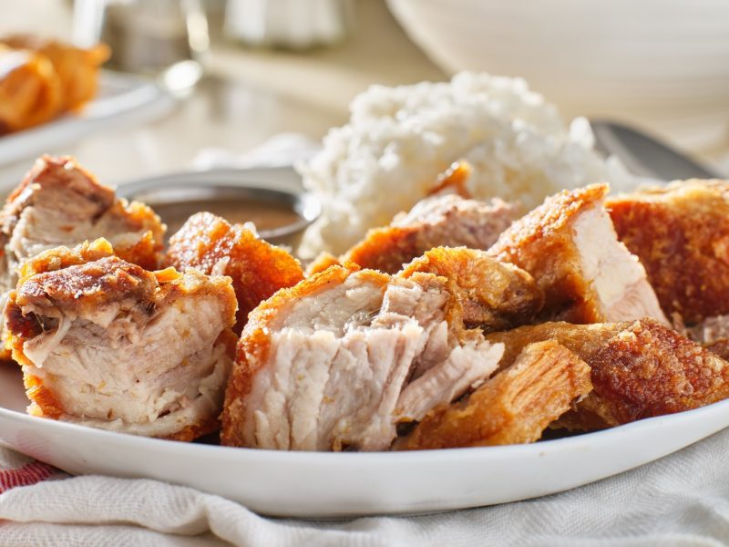 Draufsicht, leicht schräg: Ein heller Teller mit knusprig gebratenem und in Stücke geschnittenem Schweinefleisch. neben dem ist heller Reis angerichtet. Auf dem Teller steht zudem eine kleine Schale aus Metall, in der eine Sauce ist.