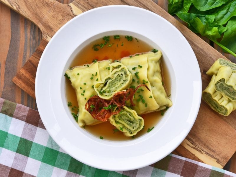 Draufsicht: Ein weißer Teller mit einer Maultaschensuppe mit Spinat auf einem Holzbrett. Drum herum liegen frische Spinatbätter, ein Küchenhandtuch und zwei angeschnittene Maultaschen.