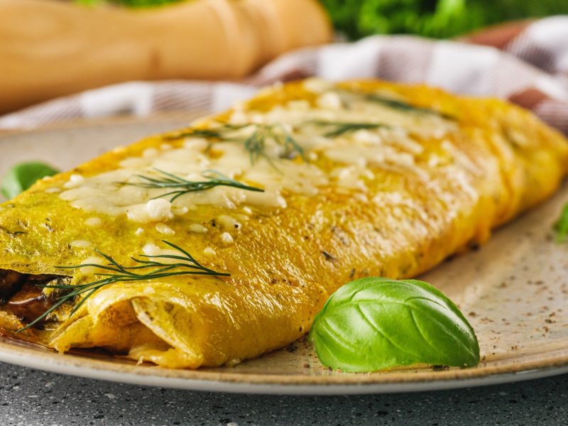 Frontal Ein französisches Omelette mit frischen Kräutern, dass zusammen geklappt ist, auf einem Teller. Im Hintergrund liegt eine lange Pfeffermühle aus Holz.