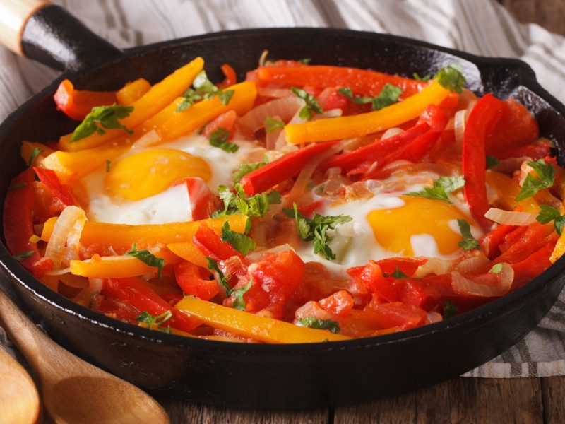 Draufsicht, schräg oben: Eine gusseiserne Pfanne mit Gemüse, wie Paprika, Tomaten und Zwiebeln. In der Mitte sind zwei Spiegeleier.