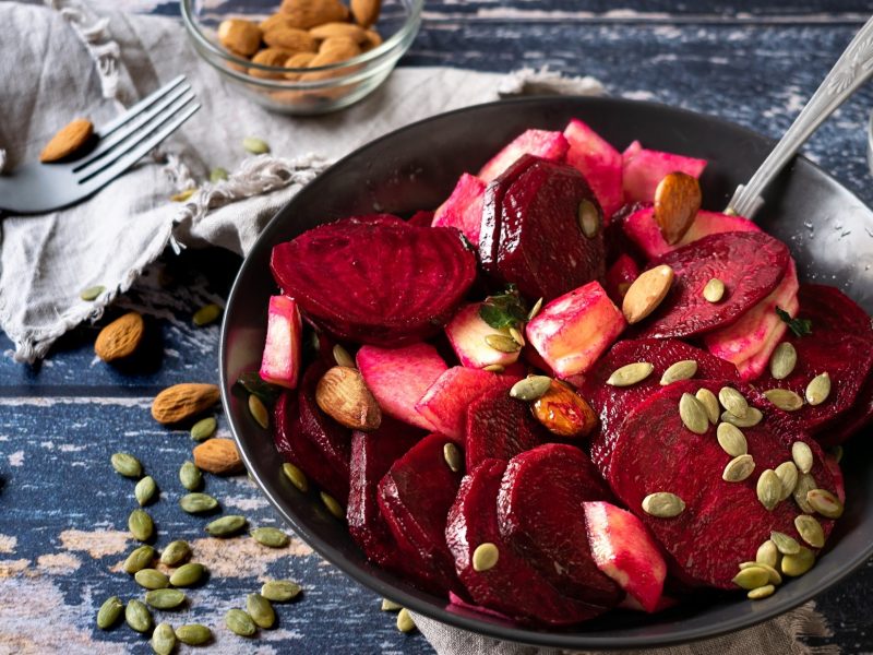 Draufsicht: Ein heller Untergrund auf dem einen Schüssel mit einem Rote-Bete-Salat mit Apfel steht, in dem ein Löffel steckt. Drum herum liegen mandeln mit Haut sowie Kürbiskerne.