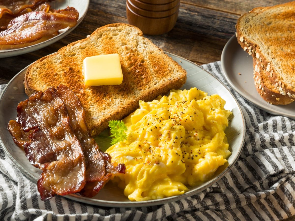 Draufsicht: Ein gedeckter Tisch. Ein großer runder Teller mit Rührei und Speck und einem Toast auf dem Butter liegt. Drumherum ein weiterer Teller mit Brot und noch ein Teller mit gebraten Speckstreifen.