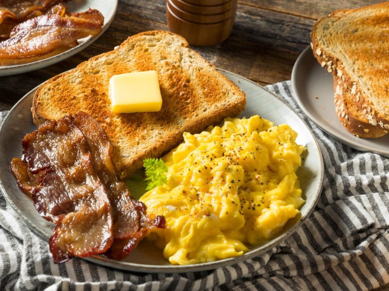 Draufsicht: Ein gedeckter Tisch. Ein großer runder Teller mit Rührei und Speck und einem Toast auf dem Butter liegt. Drumherum ein weiterer Teller mit Brot und noch ein Teller mit gebraten Speckstreifen.