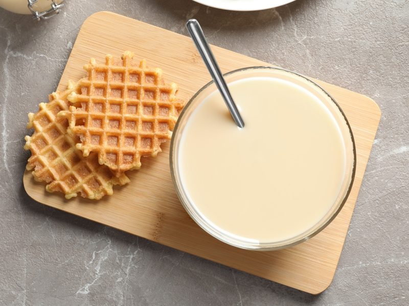 Ein kleines Schneidebrett mit der Waffelmasse und frisch ausgebackenen Waffeln auf grauem Untergrund. Alles direkt von oben fotografiert.