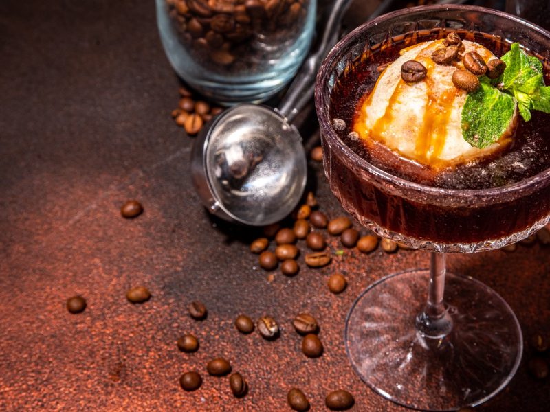 Nahaufnahme eines Glases mit cremigem Espresso Martini Affogato, dekoriert mit Karamellsirup, frischer Minze und Kaffeebohnen, umgeben von verstreuten Bohnen.