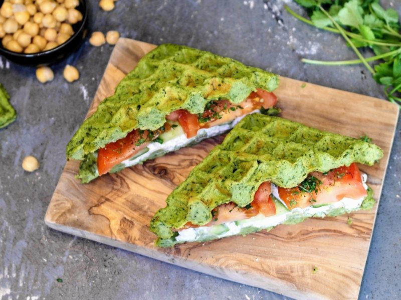 Ein kleines Holzbrett mit dem Falafel-Waffel-Sandwich auf einem steinernen Tisch. Ebenso oben im Bild sind frische Kräuter und Kichererbsen. Alles von schräg oben fotografiert.