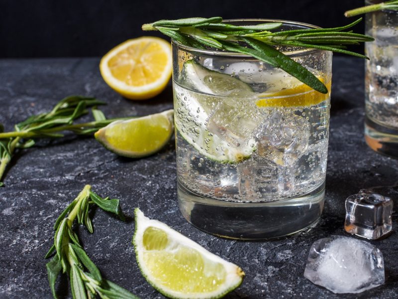 Zwei Gläser Gin Tonic mit Eis, Zitronen- und Limettenstücken, umgeben von Rosmarin und Eiswürfeln auf dunkler Steinfläche.