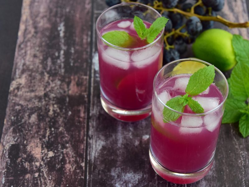 Zwei Gläser Grape Pineapple Mint Fizz mit Eiswürfeln und Minzblättern stehen auf einer rustikalen Holzoberfläche, daneben Limette und Trauben.