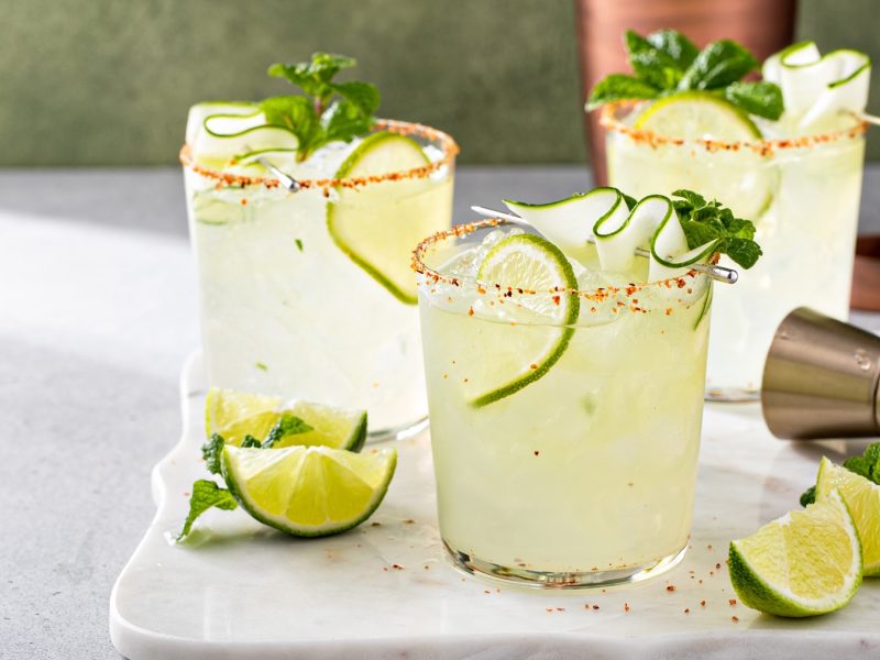 Drei Gläser mit Gurken-margaritas mit Tajin-eand auf einer weißen Marmorplatte, danebebn frische Limettenschnitze, Minze und ein Jigger.