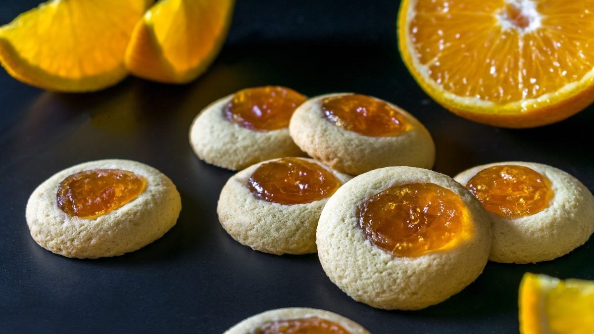 Mehrere Kekse auf einem schwarzen Untergrund mit frischen Orangenspalten und einer halbierten Orange. Alles nah und von der Seite fotografiert.