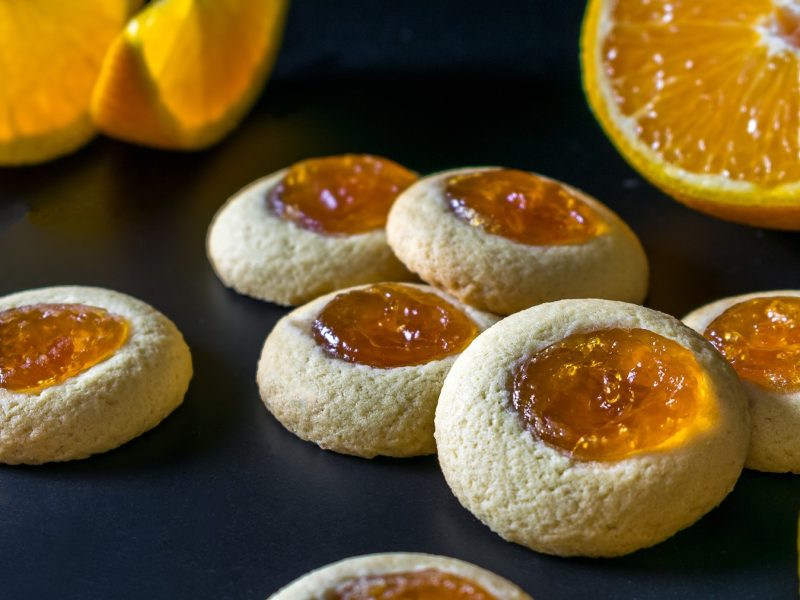 Mehrere Kekse auf einem schwarzen Untergrund mit frischen Orangenspalten und einer halbierten Orange. Alles nah und von der Seite fotografiert.