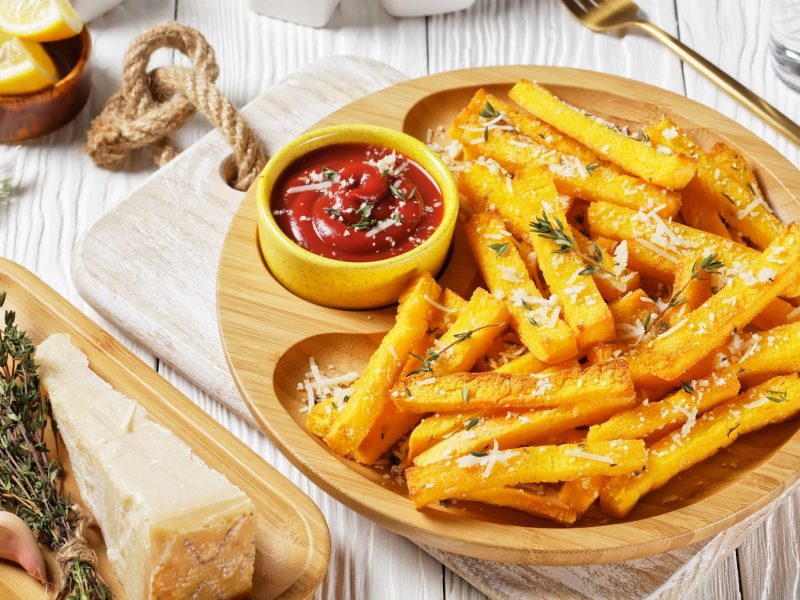 Ein Holzteller mit den Pommes auf einem hellen Tisch mit Kräutern, Parmesan, Gabel, Tuch und weiterer Dekoration. Alles von schräg oben fotografiert.