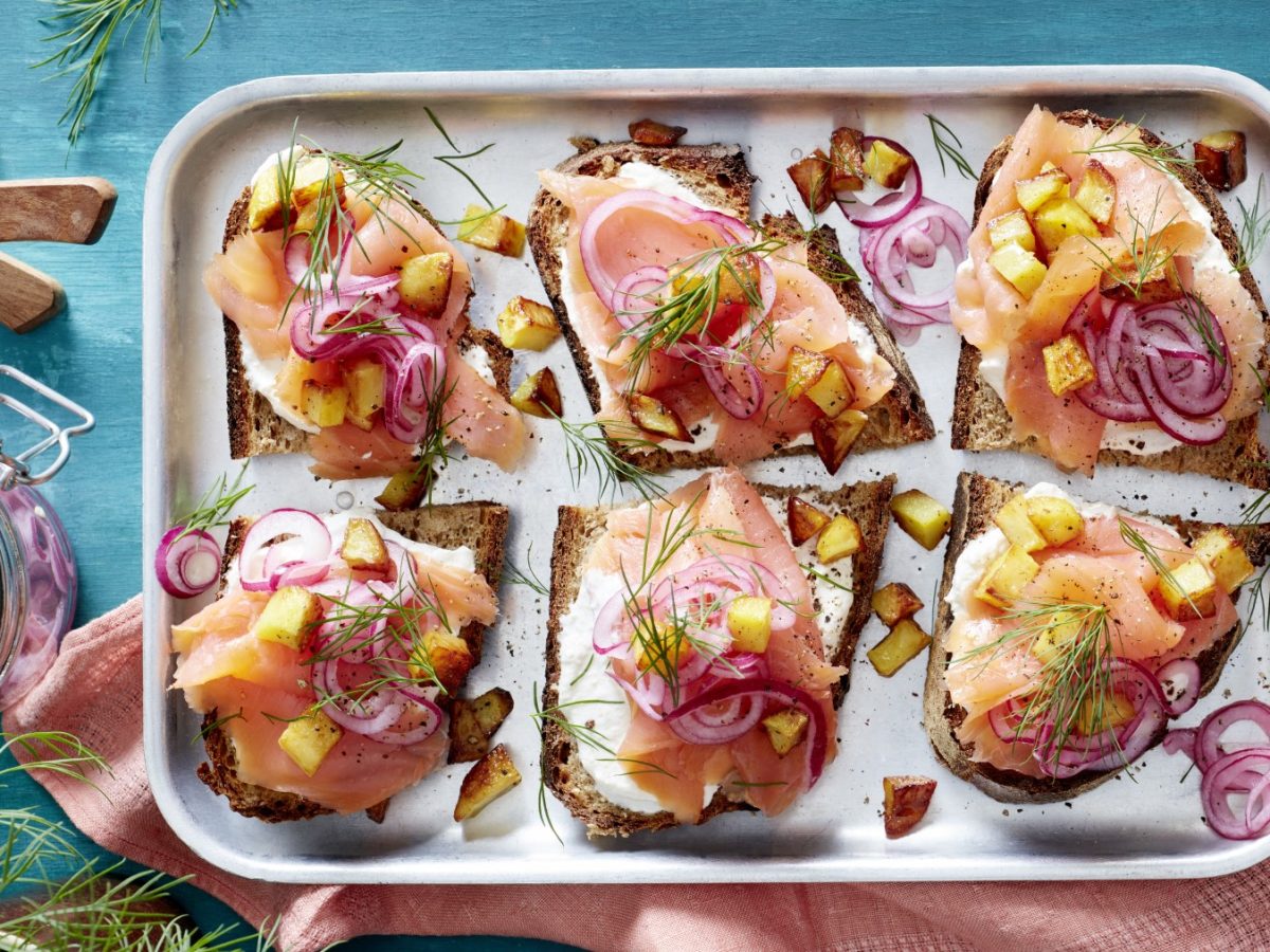 Lachs-Smörrebröd auf einem silbernen Teller auf blauem Hintergrund. Links ein Schälchen mit Zwiebeln, Messer und Dill.
