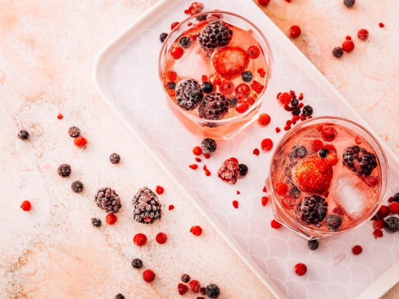Zwei Gläser mit rosa Lillet Wild Berry, garniert mit Erdbeeren, Brombeeren und roten Johannisbeeren, auf hellem Tablett.