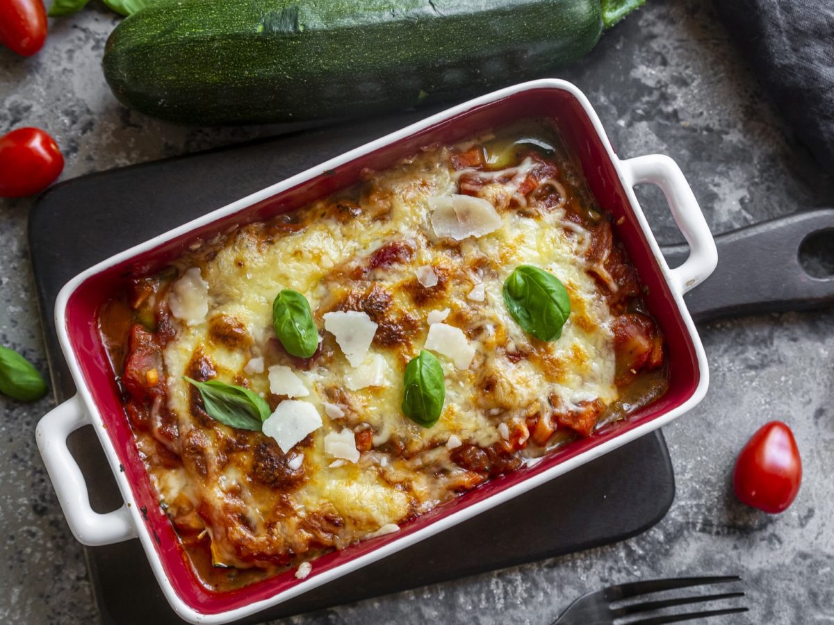 Draufsicht: eine vegetarische Lasagne mit Zucchini in einer weißen Form au einem grauen Untergrund. Drum herum um die Low-Carb-Lasagne liegen frische Basilikumblätter, eine große Zucchini und kleine Tomaten.