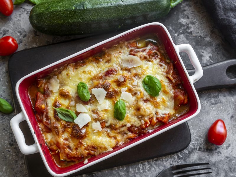 Draufsicht: eine vegetarische Lasagne mit Zucchini in einer weißen Form au einem grauen Untergrund. Drum herum um die Low-Carb-Lasagne liegen frische Basilikumblätter, eine große Zucchini und kleine Tomaten.