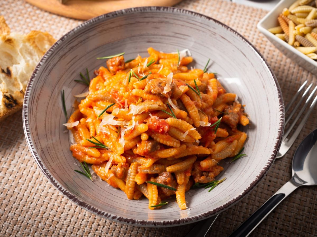 Rustikale Malloreddus alla campidanese mit kräftiger Tomatensauce, garniert mit Rosmarin und geriebenem Käse, daneben gegrilltes Brot.