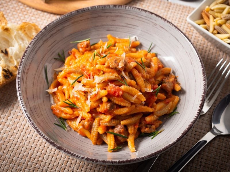 Rustikale Malloreddus alla campidanese mit kräftiger Tomatensauce, garniert mit Rosmarin und geriebenem Käse, daneben gegrilltes Brot.