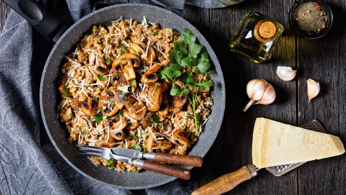 Miso-Pilzrisotto in einem grauen Teller auf dunklem Hintergrund. Links ein graues Geschirrtuch, rechts Knoblauch, Parmesan, Öl und Pfeffer.
