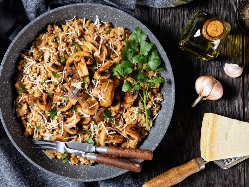 Miso-Pilzrisotto in einem grauen Teller auf dunklem Hintergrund. Links ein graues Geschirrtuch, rechts Knoblauch, Parmesan, Öl und Pfeffer.