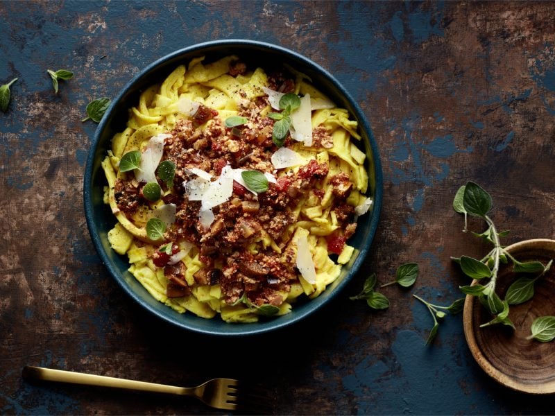Draufsicht: Schale mit Pappardelle alla Auberginen-Bolo, daneben eine Schale mit frischem Oregano auf dunklem Untergrund.