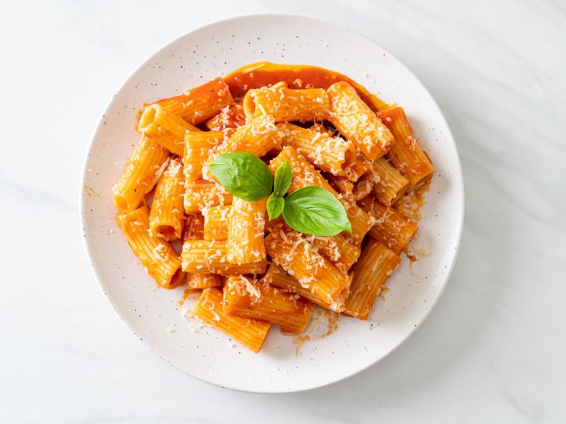 Rigatoni in leuchtend roter Paprikacreme aus dem Ofen mit geriebenem Parmesan und frischem Basilikum, serviert auf einem weißen Teller.