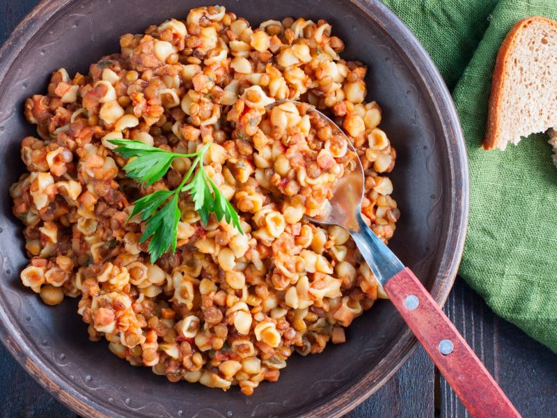 Ein brauner Teller mit kleinen Nudeln in einer würzigen Linsen-Tomatensauce "Pasta e lenticchie", garniert mit frischer Petersilie, auf dunklem Holzuntergrund.