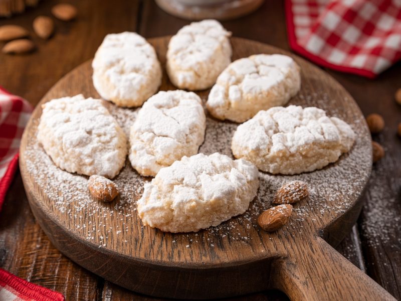 Italienische Pasta di mandorle mit Puderzucker liegen auf einem rustikalen Holzbrett, umgeben von ganzen Mandeln und karierten Stoffservietten.