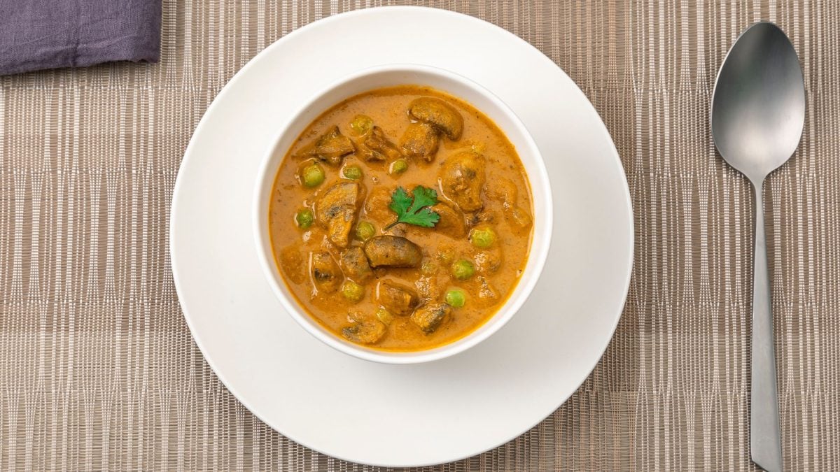 Würziges Pilz-Curry mit Erbsen in einer weißen Schüssel, serviert auf einem gedeckten Tisch mit Löffel und Stoffserviette.