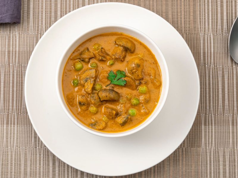 Würziges Pilz-Curry mit Erbsen in einer weißen Schüssel, serviert auf einem gedeckten Tisch mit Löffel und Stoffserviette.